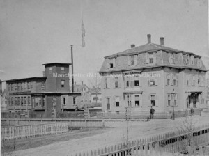 View of the Corey Needle Works on Concord Street. Looking south east Maple Street is seen in the foreground and runs north and south. A man stands on the corner in front of a three story second empire house with mansard roof. [That house is William Corey's home, and it is still standing].